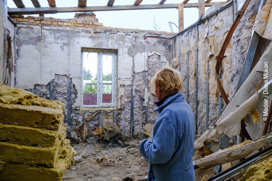 Eine Person blickt in das zerstörte Innere eines Hauses mit freigelegten Wänden – sichtbare Folgen des Ukrainekriegs und der dringende Bedarf an Wiederaufbauhilfe.