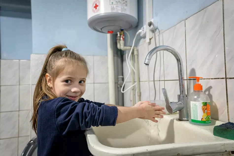 Ein Mädchen wäscht sich lächelnd die Hände an einem Wasserhahn – Zugang zu sauberem Wasser und Hygiene dank humanitärer Hilfe in der Ukraine.