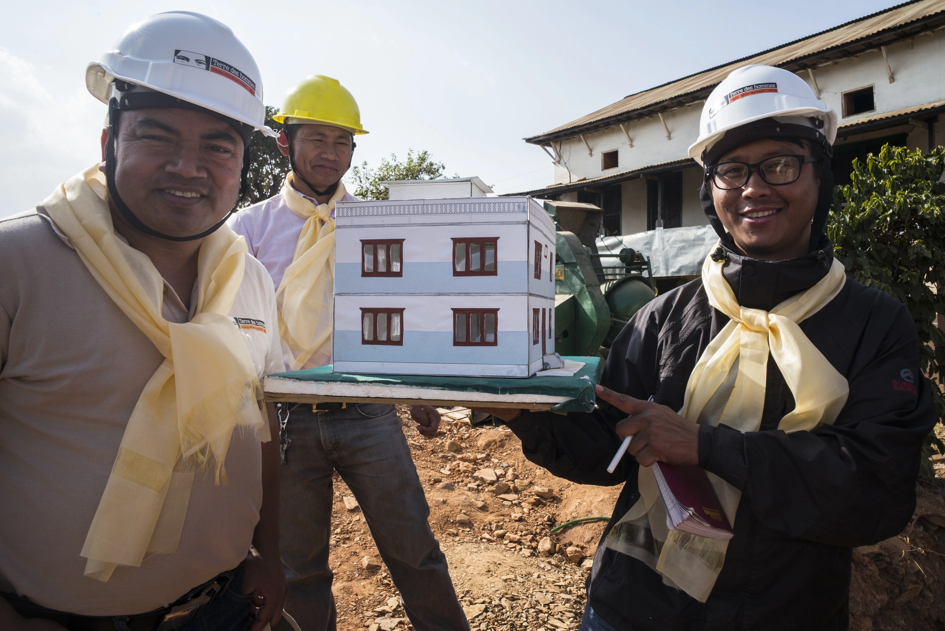 Drei Männer mit Schutzhelmen zeigen ein Modellhaus für sicheren Wiederaufbau nach Katastrophen zur Stärkung von Resilienz und Schutz.