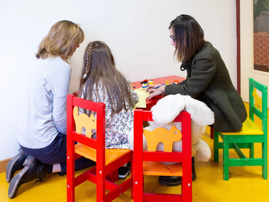 Ein Mädchen sitzt mit zwei erwachsenen Frauen an einem kleinen Tisch, alle mit dem Rücken zur Kamera.