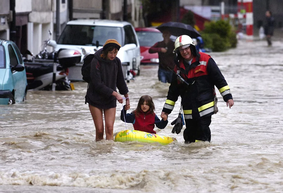 Ein Mann und eine Frau halten ein Kind an der Hand, das auf einem gelben Schlauchboot sitzt und durch die überfluteten Straßen läuft, während die Schweizer Hochwasserhilfe 2005 im Einsatz ist.