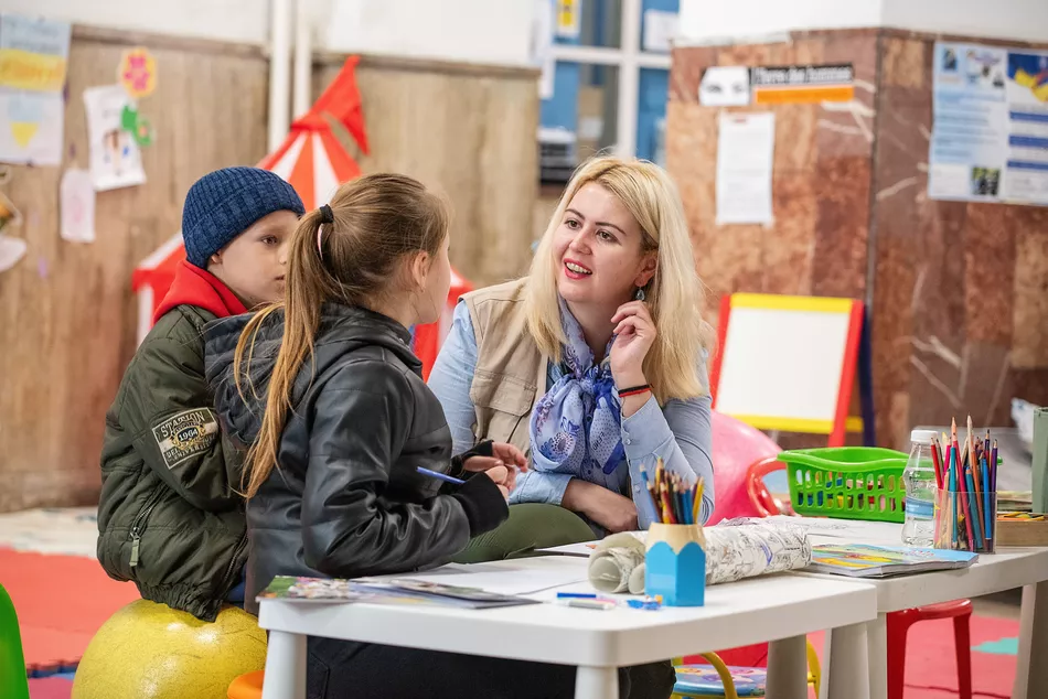 Eine Lehrkraft spricht mit zwei Kindern in einem bunt eingerichteten Lernraum – Bildungsangebote für geflüchtete Kinder in der Ukraine helfen, trotz Krieg weiter zu lernen.